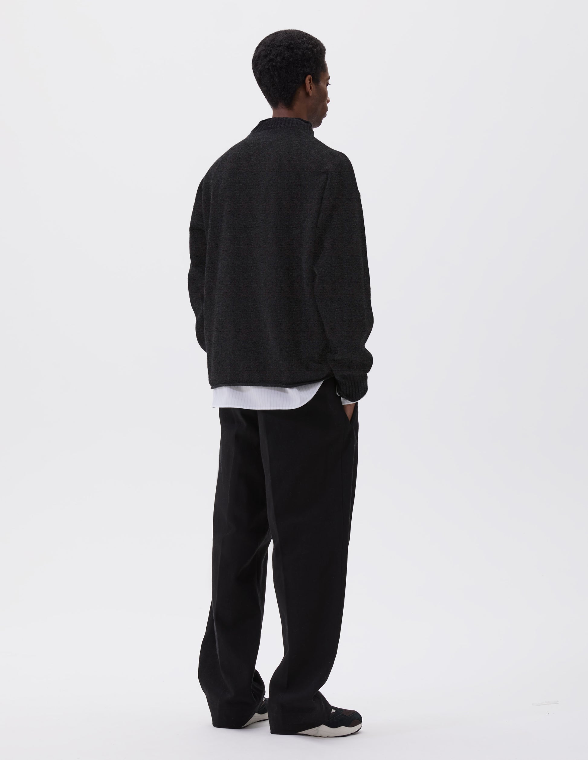 Full body image, back view of model wearing black long sleeve jumper with rolled edge neck and hem and contrasting rib on neck and sleeves. Styled with black trousers, white and grey striped shirt and black and brown trainers.