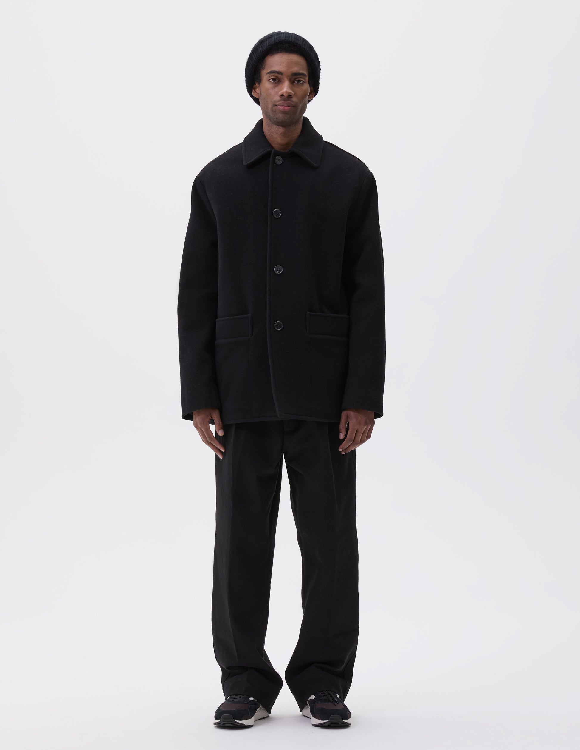 Full body image, front view of model wearing black wool coat with contrast wool cotton fleece lining, with deep relaxed welt pockets and stitch through detailing. Finished with horn button closure. Styled with grey hat, black jumper, black trousers and black and brown trainers. 