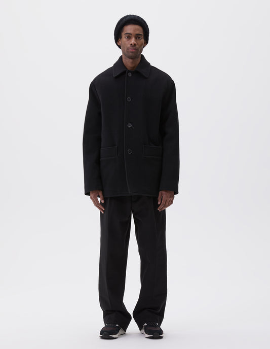 Full body image, front view of model wearing black wool coat with contrast wool cotton fleece lining, with deep relaxed welt pockets and stitch through detailing. Finished with horn button closure. Styled with grey hat, black jumper, black trousers and black and brown trainers. 