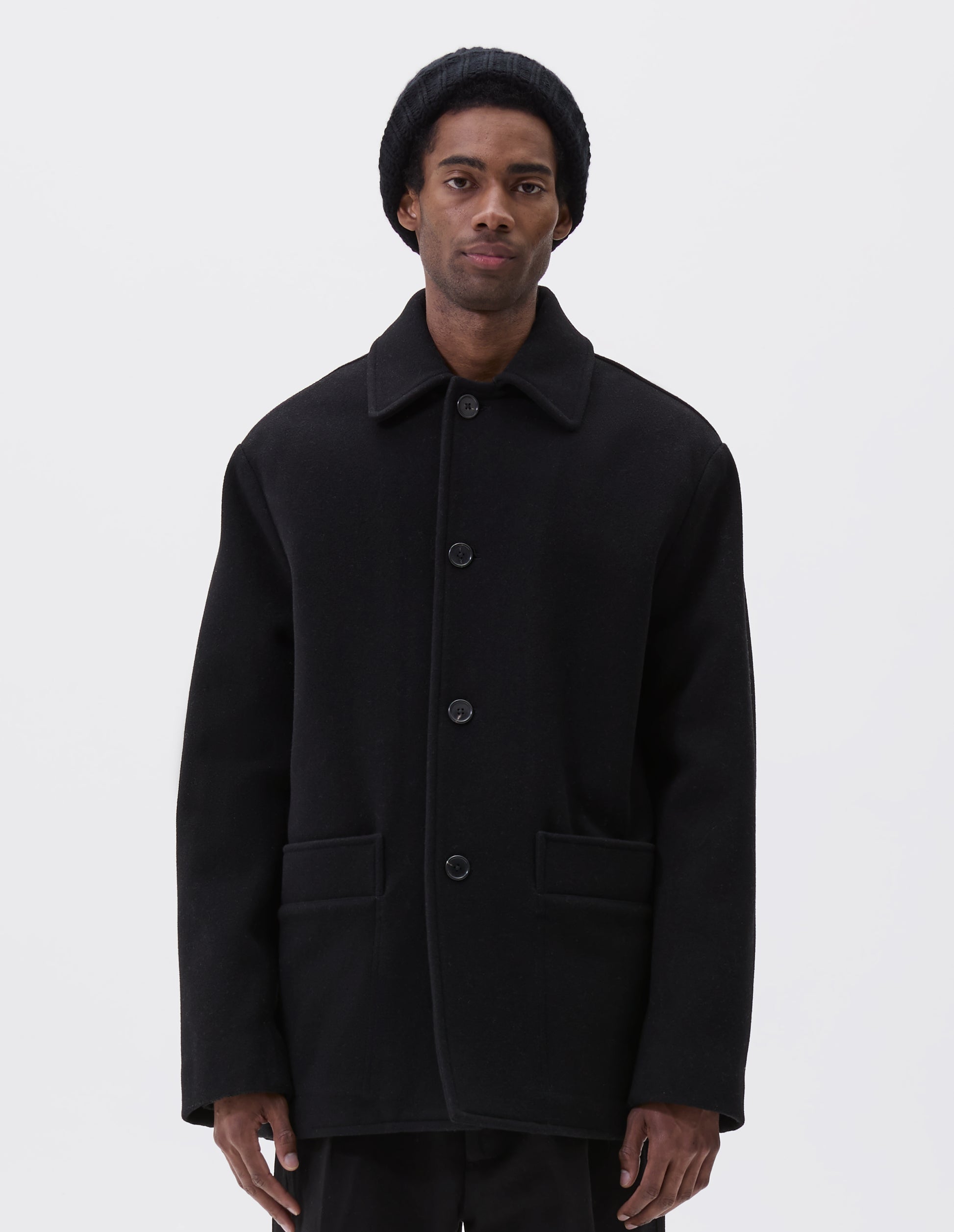 Cropped body image, front view of model wearing black wool coat with contrast wool cotton fleece lining, with deep relaxed welt pockets and stitch through detailing. Finished with horn button closure. Styled with grey hat, black jumper and black trousers.