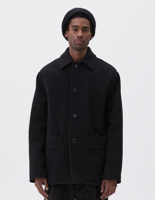 Cropped body image, front view of model wearing black wool coat with contrast wool cotton fleece lining, with deep relaxed welt pockets and stitch through detailing. Finished with horn button closure. Styled with grey hat, black jumper and black trousers.