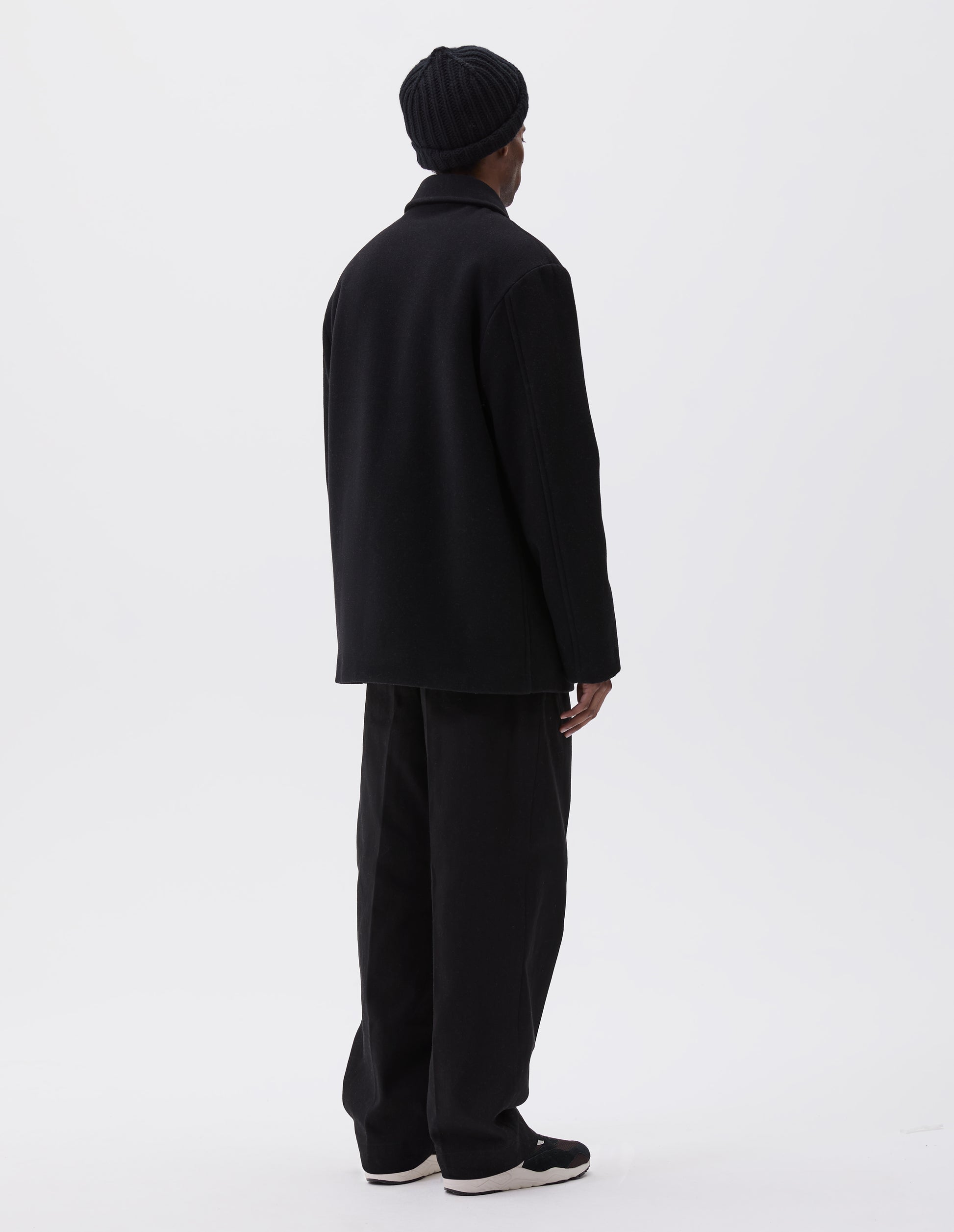 Full body image, back view of model wearing black wool coat with contrast wool cotton fleece lining, with deep relaxed welt pockets and stitch through detailing. Styled with grey hat, black trousers and black and brown trainers.