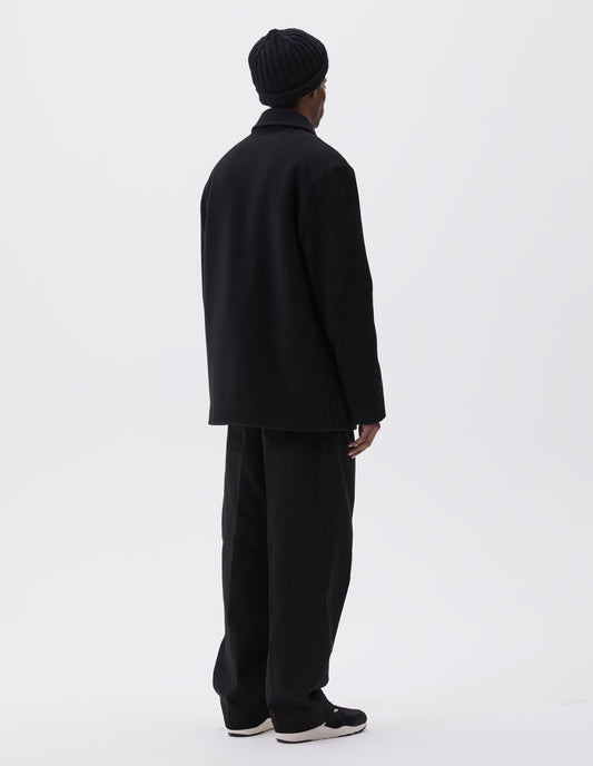 Full body image, back view of model wearing black wool coat with contrast wool cotton fleece lining, with deep relaxed welt pockets and stitch through detailing. Styled with grey hat, black trousers and black and brown trainers.