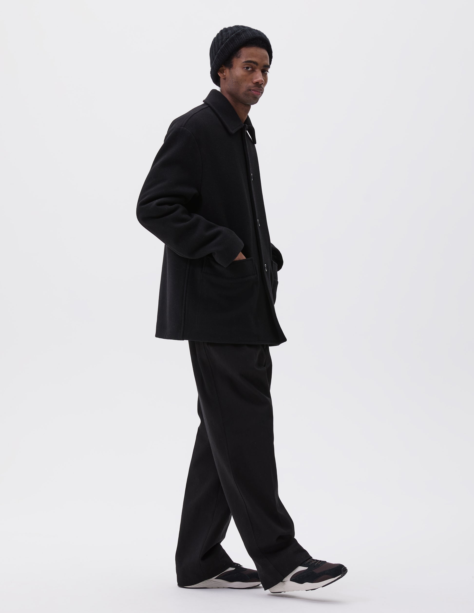 Full body image, side view of model wearing black wool coat with contrast wool cotton fleece lining, with deep relaxed welt pockets and stitch through detailing. Finished with horn button closure. Styled with grey hat, black jumper, black trousers, and black and brown trainers. 