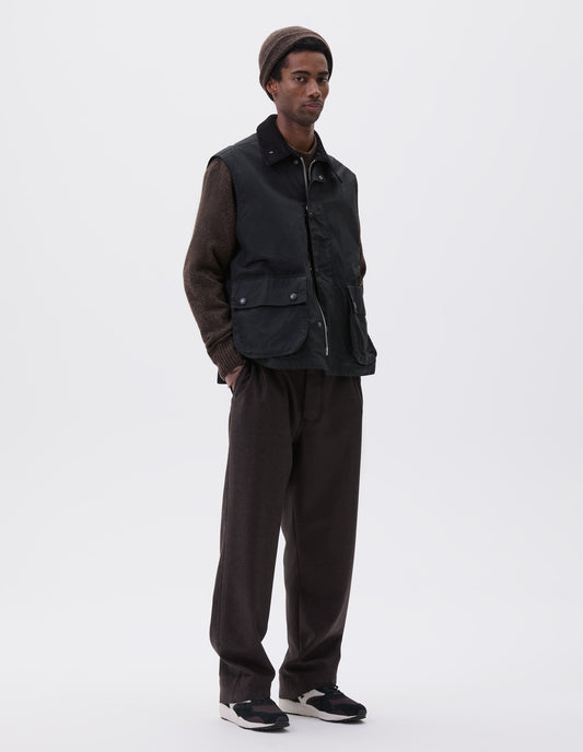 Full body image, front view of model wearing black relaxed gilet with storm tab on corduroy collar, two-way zip fastening and press stud fastening storm flap, large bellows pockets with press stud fastening envelope closure. Styled over brown jumper with brown beanie, brown trousers and black, brown and white trainers. 
