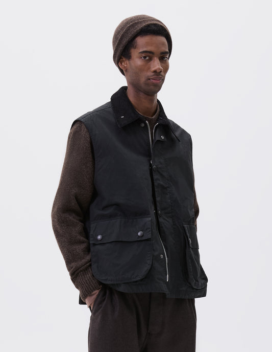 Cropped image, front view of model wearing black relaxed gilet with storm tab on corduroy collar, two-way zip fastening and press stud fastening storm flap, large bellows pockets with press stud fastening envelope closure. Styled over brown jumper with brown beanie and brown trousers. 
