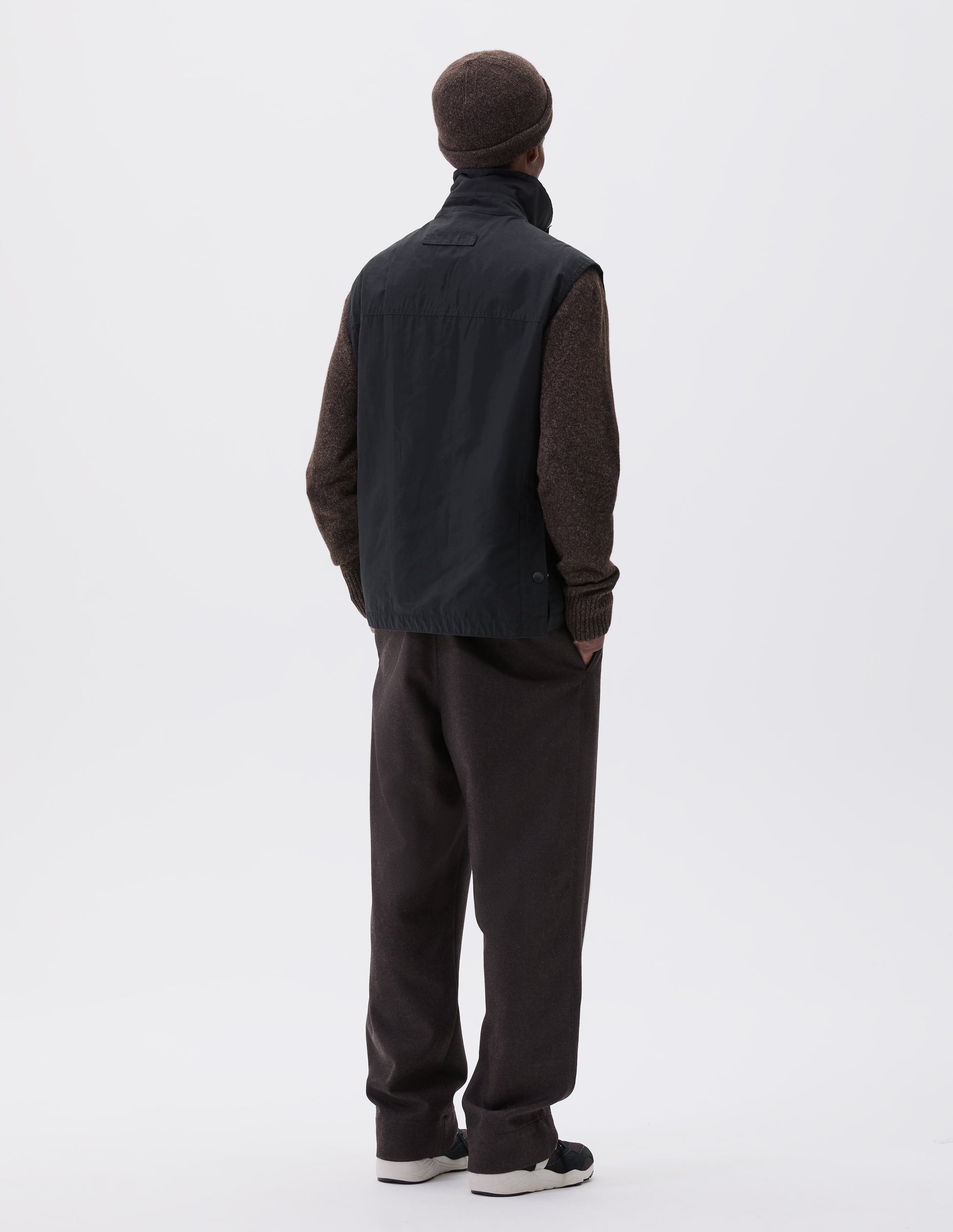 Full body image, back view of model wearing black relaxed gilet with stand collar, hanger loop on back and press stud fastening vent split hem with gussets. Styled over brown jumper with brown beanie, brown trousers and black, brown and white trainers.