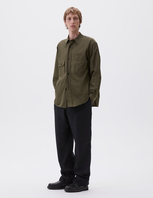 Full body image, front view of model wearing green shirt with patch pocket and button-through flap pocket on chest, raised placket with horn buttons, styled with dark blue trousers and black leather shoes. 