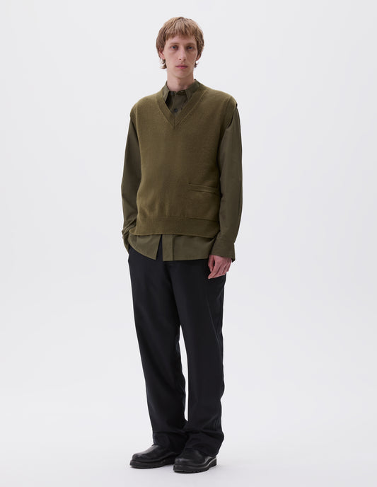 Full body image, front view of model wearing olive green sleeveless knitted jumper, with contrast rib on neck, armholes and hem, with pocket on lower hip. Styled with green brown shirt, navy trousers and black leather boots.