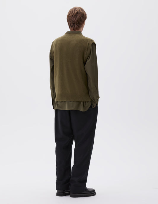 Full body image, back view of model wearing olive green sleeveless knitted jumper, with contrast rib on neck, armholes and hem. Styled with green brown shirt, navy trousers and black leather boots.