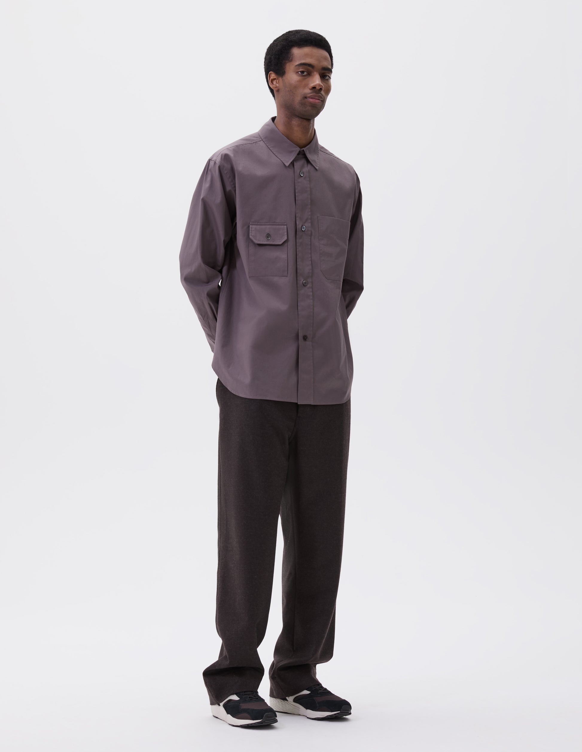 Full body image, front view of model wearing grey shirt with patch pocket and button-through flap pocket on chest, raised placket with mother of pearl buttons, styled with dark grey trousers and black, brown and white trainers.