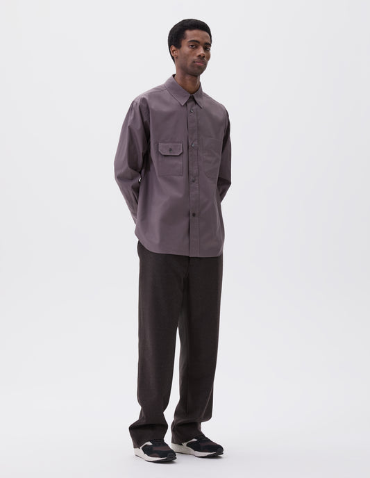 Full body image, front view of model wearing grey shirt with patch pocket and button-through flap pocket on chest, raised placket with mother of pearl buttons, styled with dark grey trousers and black, brown and white trainers.