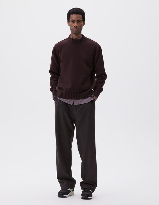 Full body image, front view of model wearing dark brown ribbed crew neck jumper with ribbed cuffs and hem, and patch pocket with rib detail, styled over grey shirt, dark grey trousers and black, brown and white trainers.
