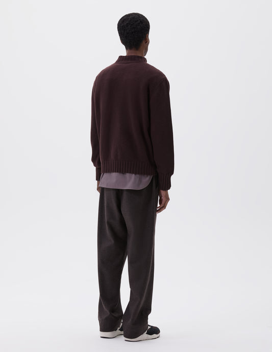 Full body image, back view of model wearing dark brown ribbed crew neck jumper with ribbed cuffs and hem, styled over grey shirt, dark grey trousers and black, brown and white trainers.
