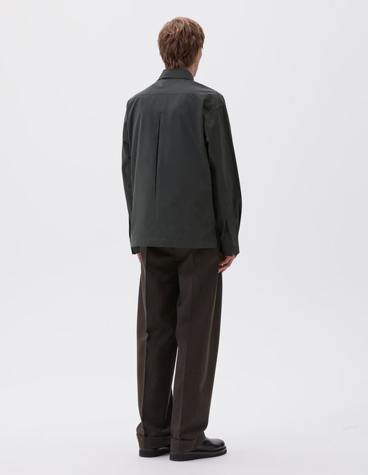 Full body image, front view of model wearing dark green shirt with vent split hem, deep inverted pleat on back yoke and horn buttons on cuffs, styled with dark brown trousers and black leather shoes.