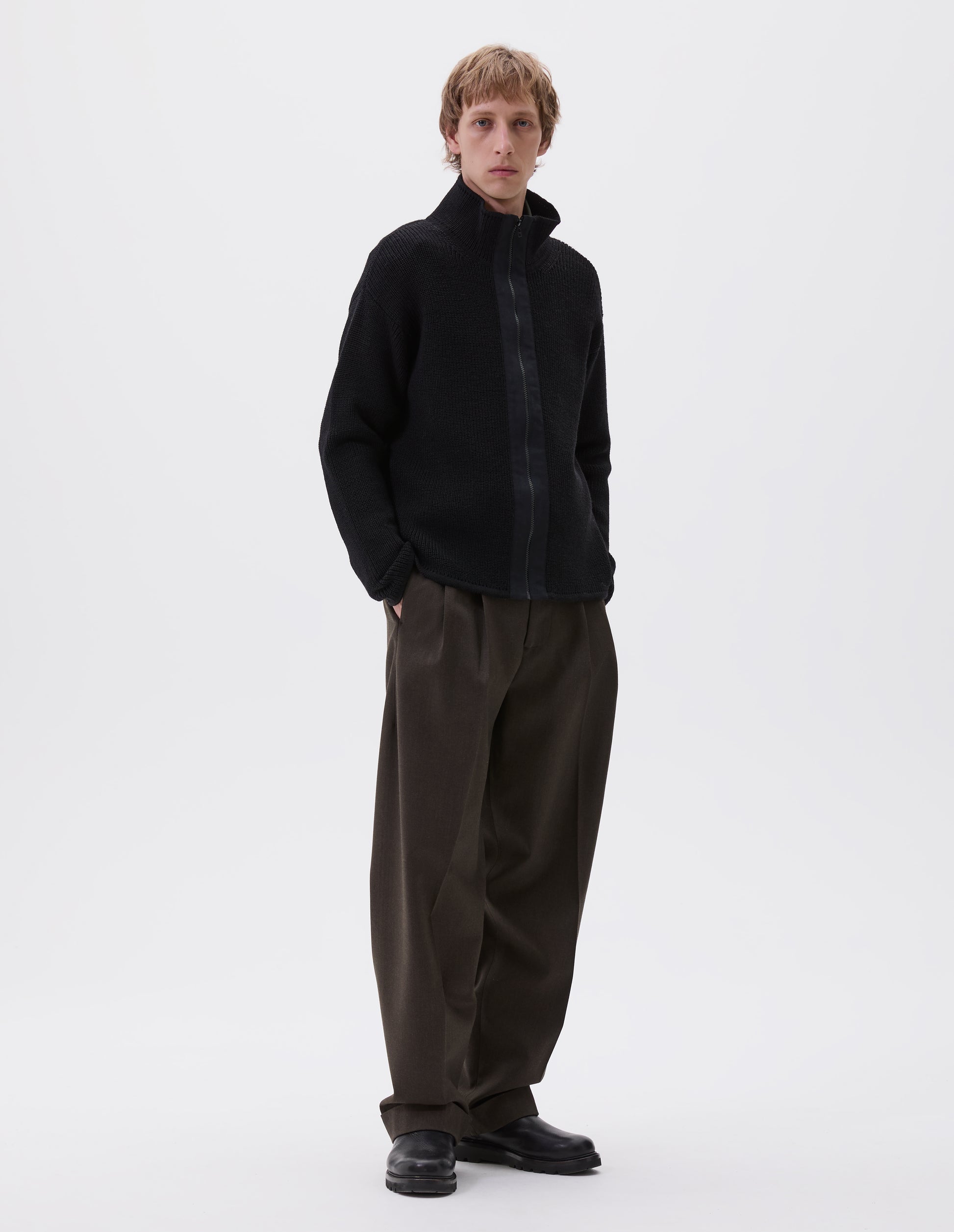 Full body image, front view of model wearing black zip through wool cardigan with stand collar, straight hem and cuffs and contrast cotton tape placket with chunky Vislon zip fastening. Styled with greenish brown trousers and black leather boots.