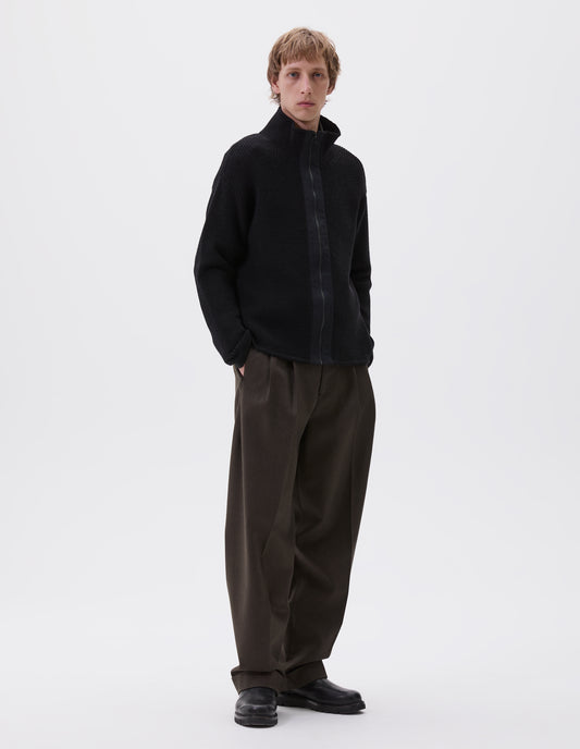 Full body image, front view of model wearing black zip through wool cardigan with stand collar, straight hem and cuffs and contrast cotton tape placket with chunky Vislon zip fastening. Styled with greenish brown trousers and black leather boots.