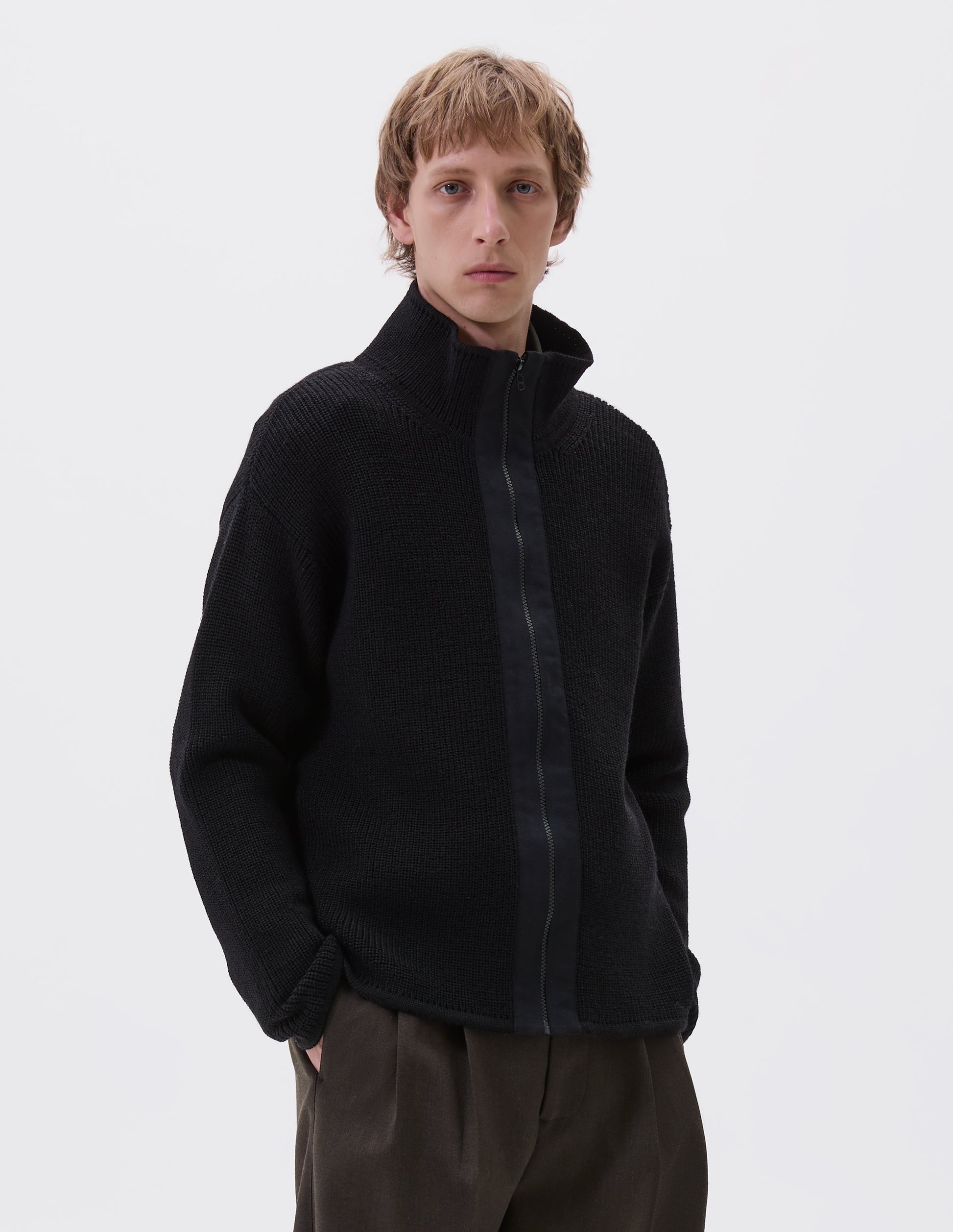 Cropped body image, front view of model wearing black zip through wool cardigan with stand collar, straight hem and cuffs and contrast cotton tape placket with chunky Vislon zip fastening. Styled with greenish brown trousers.