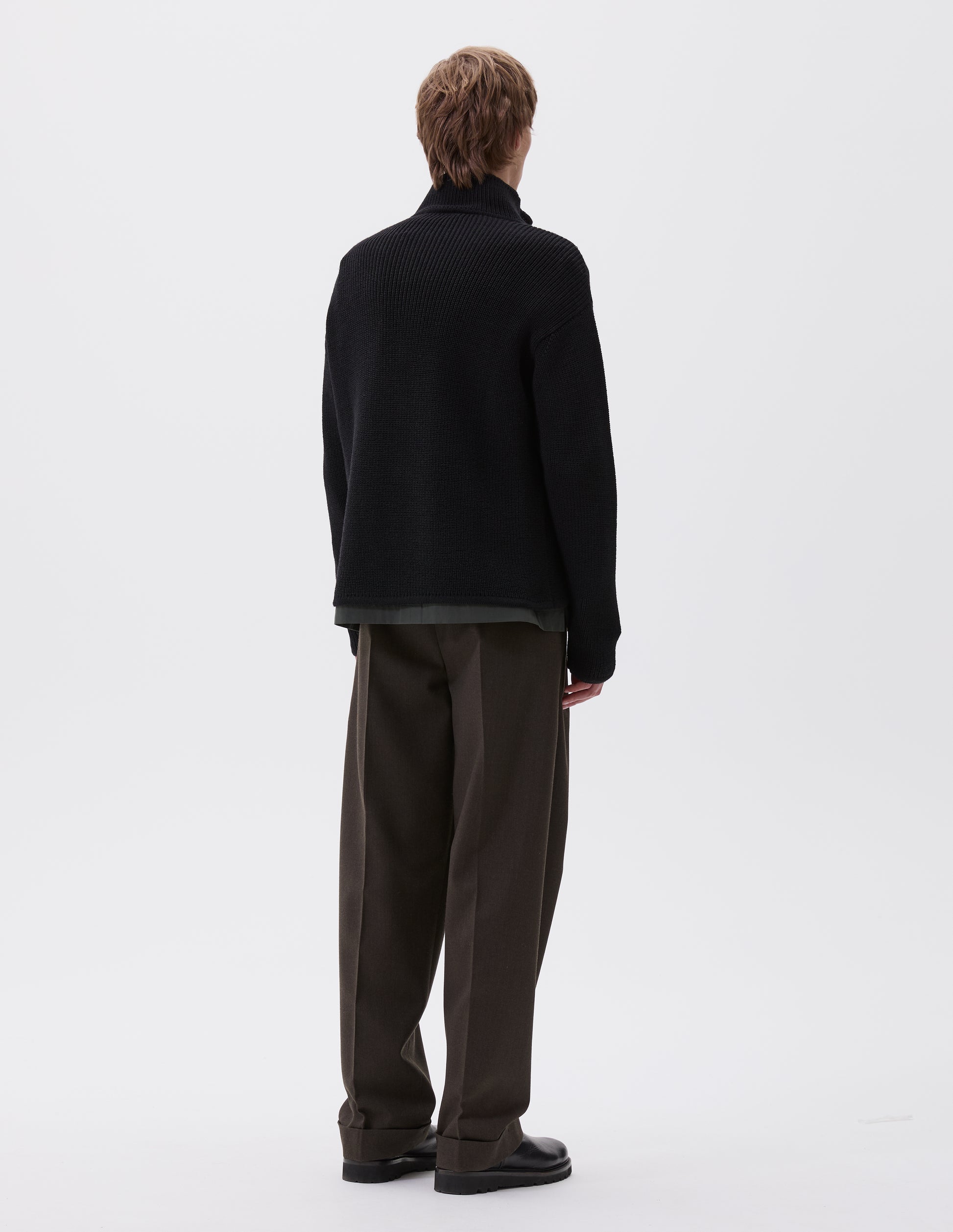 Full body image, back view of model wearing black zip through wool cardigan with straight hem and cuffs. Styled with greenish brown trousers and black leather boots.