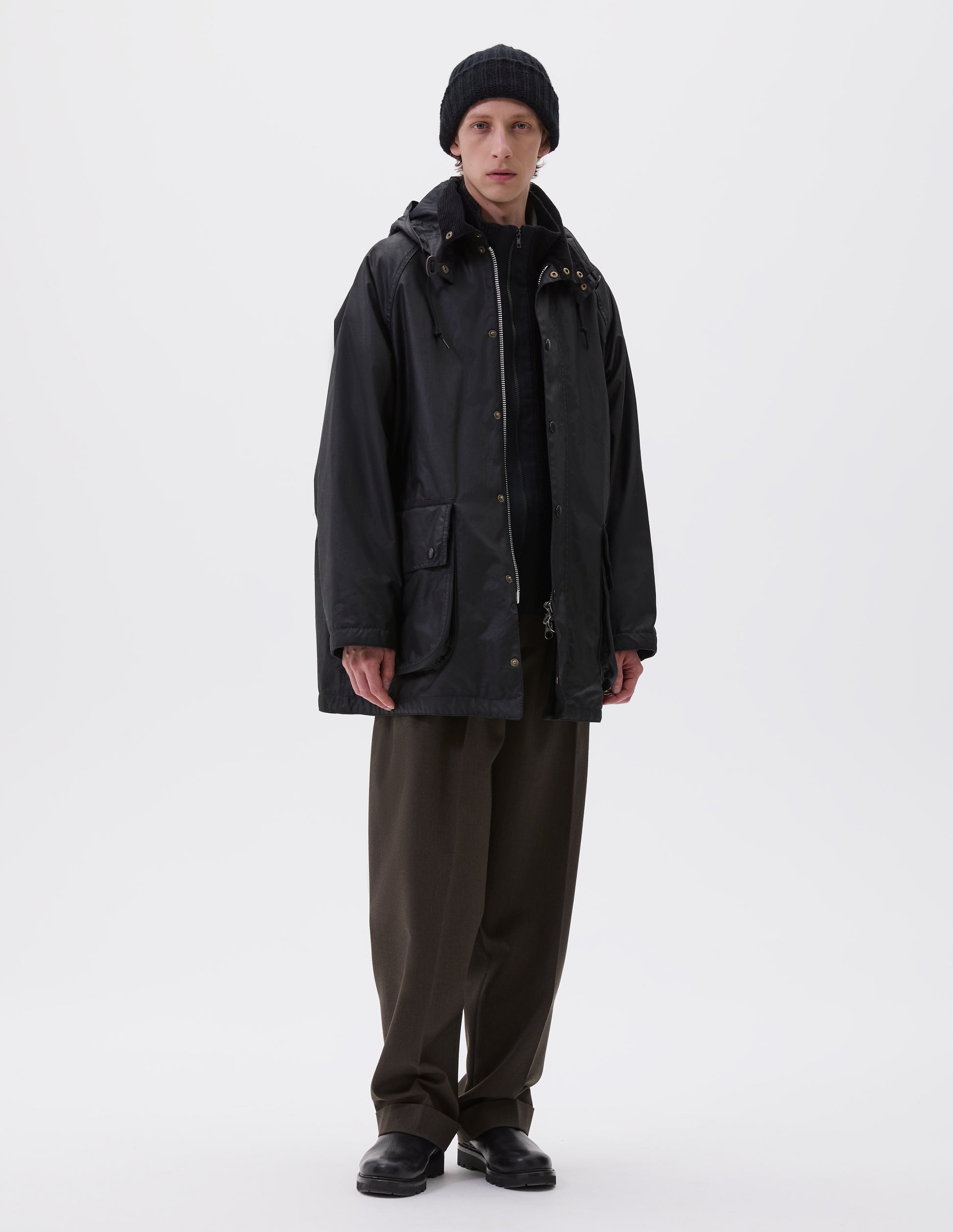 Full body image, front view of model wearing oversized black parka with corduroy collar and hood with wire adjustable peak, raglan sleeve, two way zip fastening, press stud fastening storm flap and large bellows pockets with press stud fastening envelope closure. Styled over black jumper with black beanie, brown trousers and black boots.