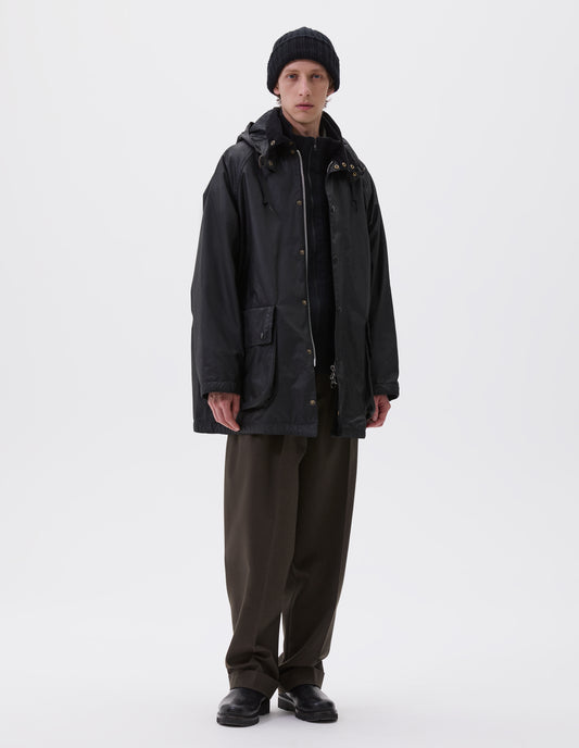 Full body image, front view of model wearing oversized black parka with corduroy collar and hood with wire adjustable peak, raglan sleeve, two way zip fastening, press stud fastening storm flap and large bellows pockets with press stud fastening envelope closure. Styled over black jumper with black beanie, brown trousers and black boots.