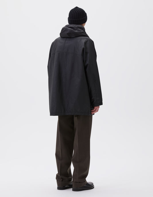 Full body image, back view of model wearing oversized black parka with hood and raglan sleeves. Styled with black beanie, brown trousers and black boots.