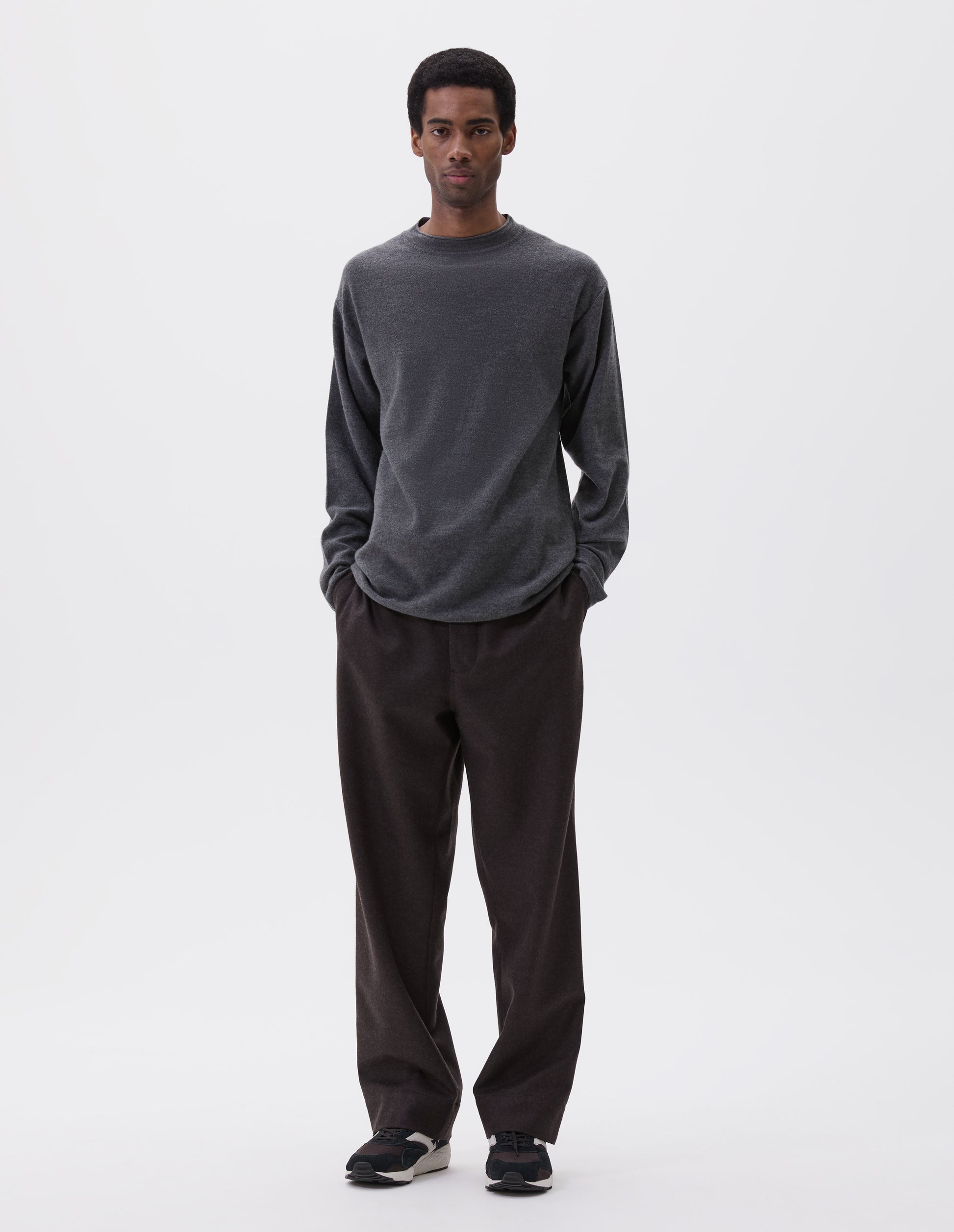 Full body image, front view of model wearing relaxed  grey crew neck jumper with rolled edge detailing on neck, rib trim on cuffs and hem. Styled with brown trousers and black, brown and white trainers.