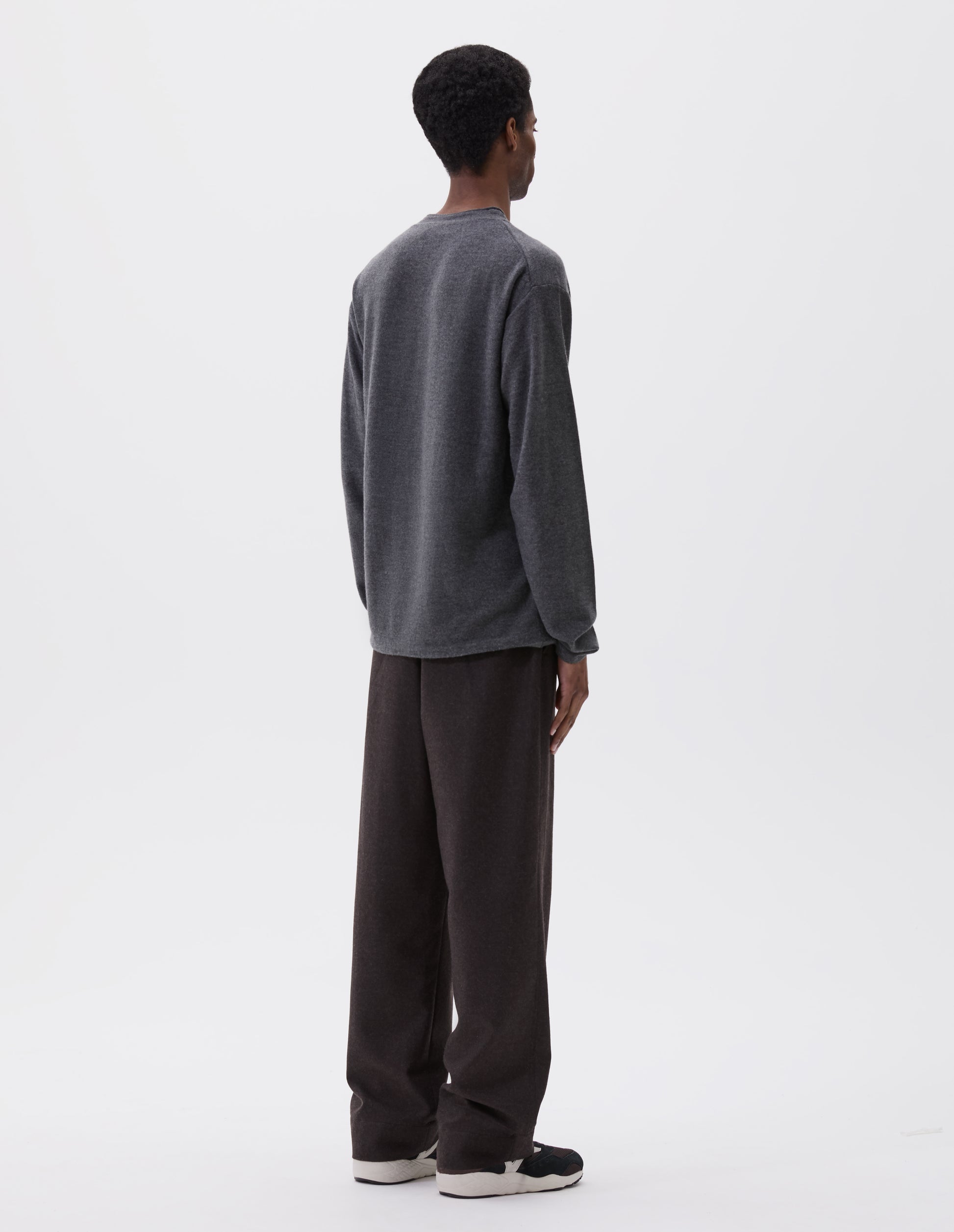 Full body image, back view of model wearing relaxed grey crew neck jumper with rolled edge detailing on neck, rib trim on cuffs and hem. Styled with brown trousers and black, brown and white trainers.