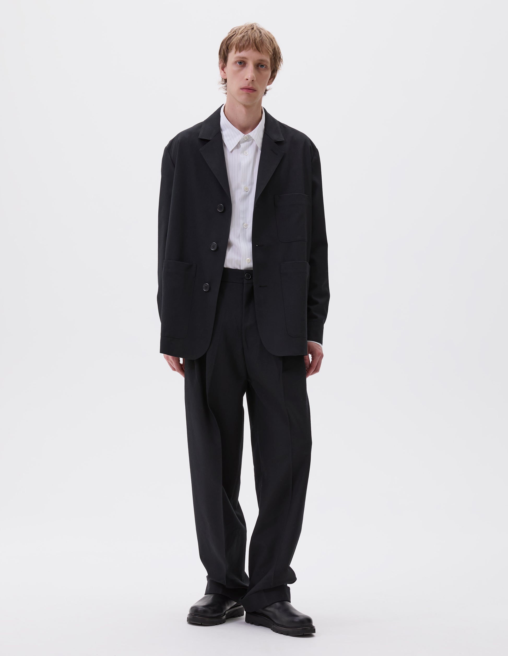 Full body image, front view of model wearing black wool blazer with horn button fastening, deep wrap closure, shirt cuff sleeves and patch pockets. Styled with white and grey striped shirt and matching black wool trousers. With black leather boots.