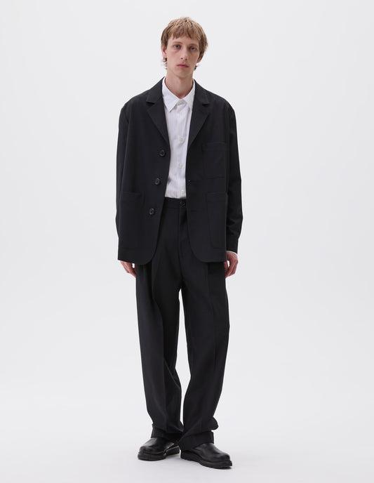 Full body image, front view of model wearing black wool blazer with horn button fastening, deep wrap closure, shirt cuff sleeves and patch pockets. Styled with white and grey striped shirt and matching black wool trousers. With black leather boots.