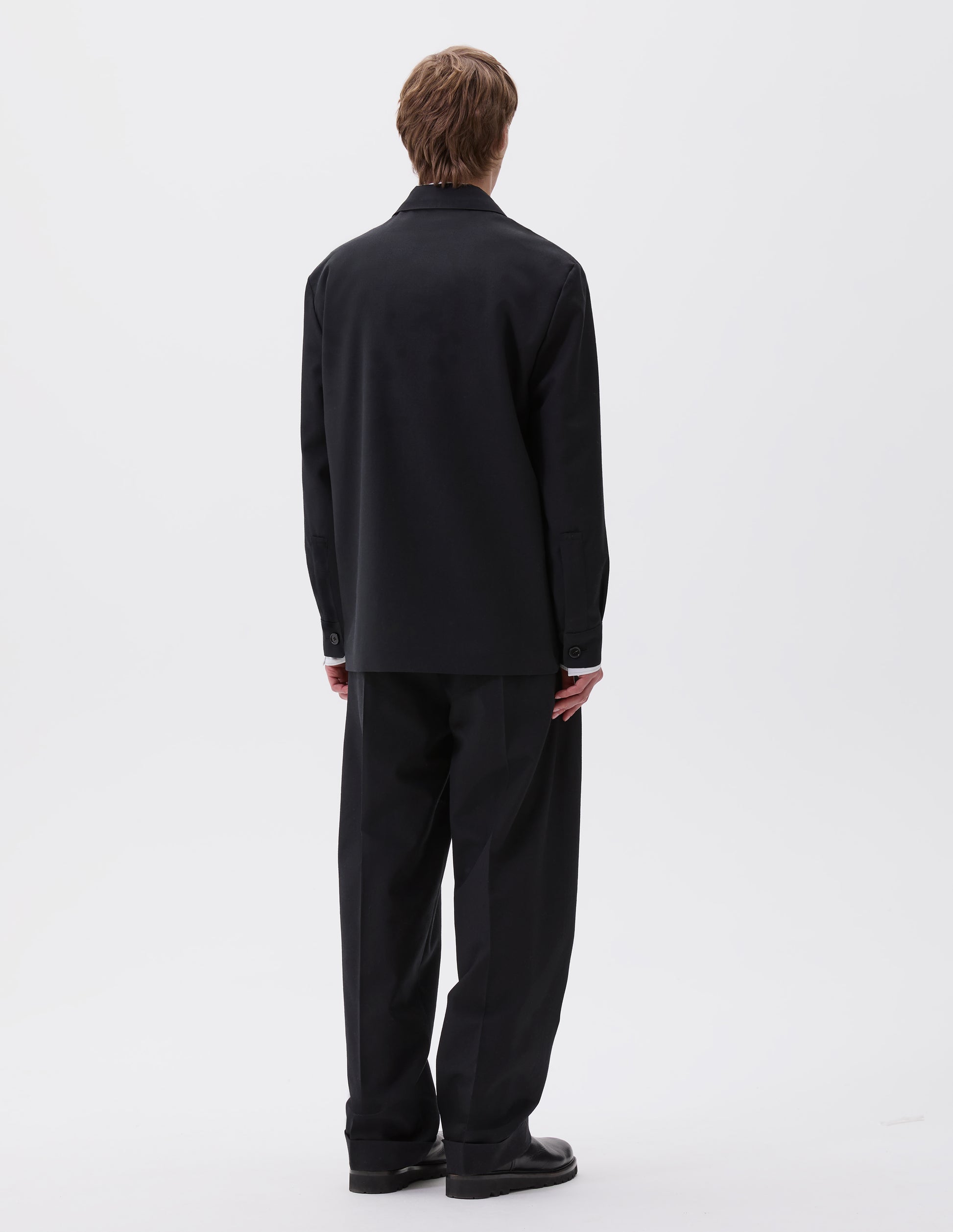 Full body image, back view of model wearing black wool blazer with horn button shirt cuffs. Styled with matching black trousers and black leather boots.