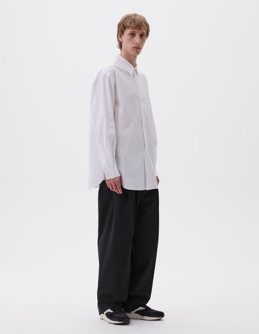 Full body image, front view of model wearing white cotton shirt with chest pocket, curved hem, curved placket from waist to hem and mother of pearl buttons. Styled with black trousers and black and brown trainers.