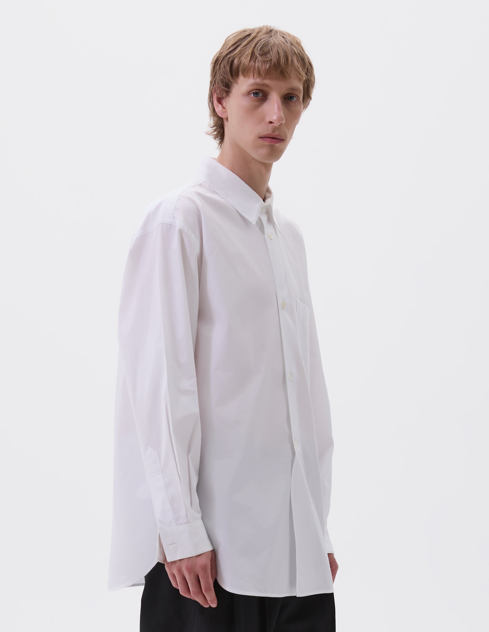 Cropped body image, front view of model wearing white cotton shirt with chest pocket, curved hem, curved placket from waist to hem and mother of pearl buttons. Styled with black trousers.
