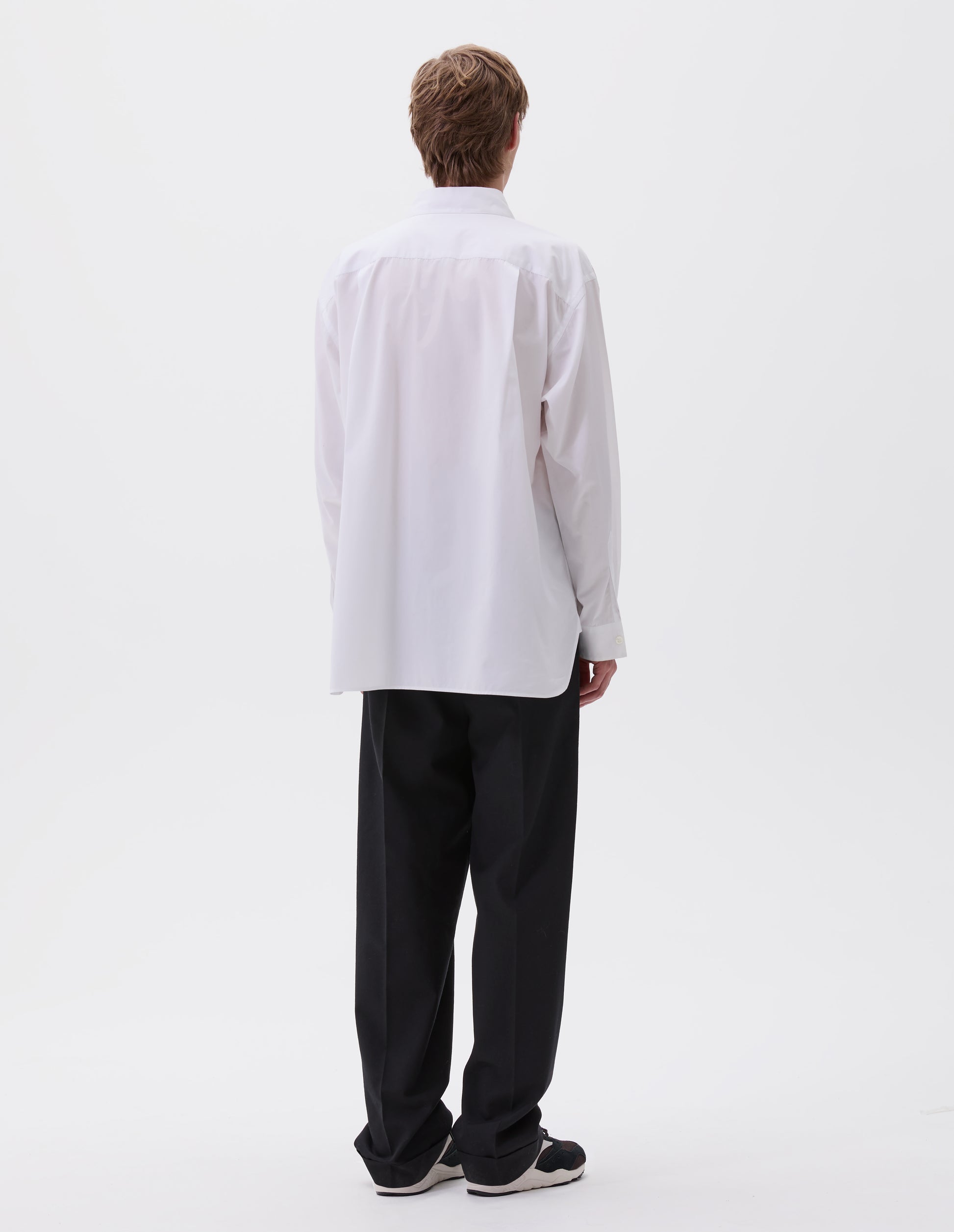 Full body image, back view of model wearing white cotton shirt, with curved hem, pleats on back yoke and mother of pearl button cuffs. Styled with black trousers and black and brown trainers.