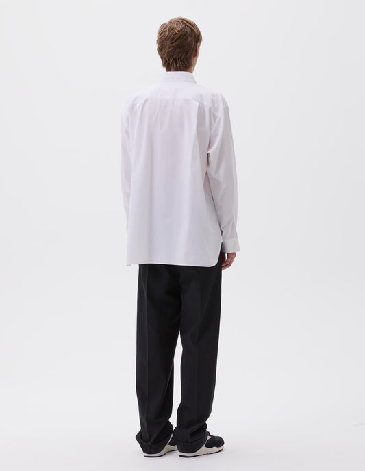 Full body image, back view of model wearing white cotton shirt, with curved hem, pleats on back yoke and mother of pearl button cuffs. Styled with black trousers and black and brown trainers.