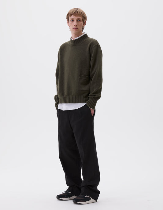 Full body image, front view of model wearing dark green ribbed crew neck jumper with ribbed cuffs and hem, and patch pocket with rib detail, styled over white striped shirt, black trousers and black, brown and white trainers.