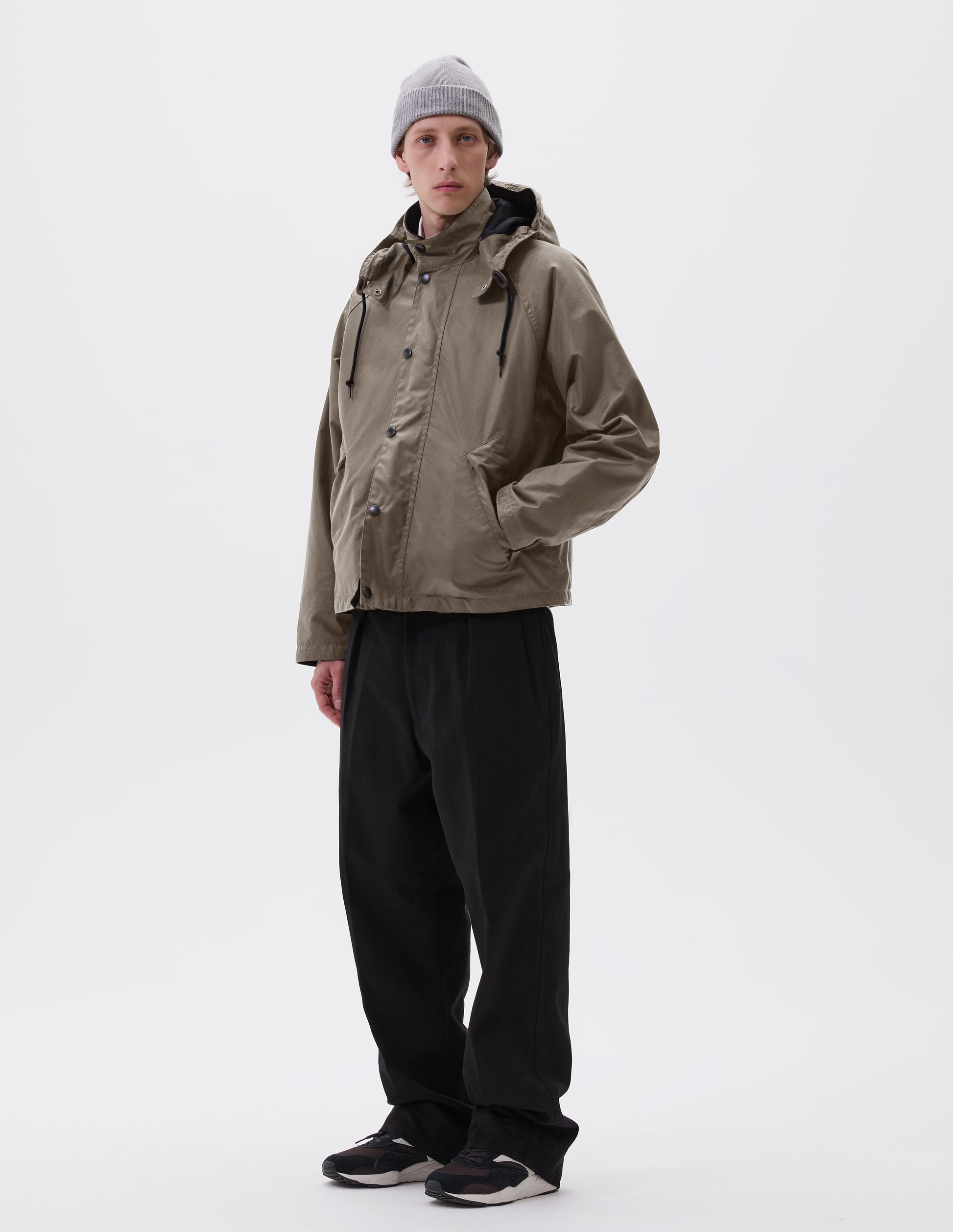Full body image, front view of model wearing boxy dark beige jacket with stand collar and hood with wire adjustable peak, raglan sleeve, press stud storm flap and wide storm welt side pockets. Styled with grey beanie, black trousers and black, brown and white trainers.