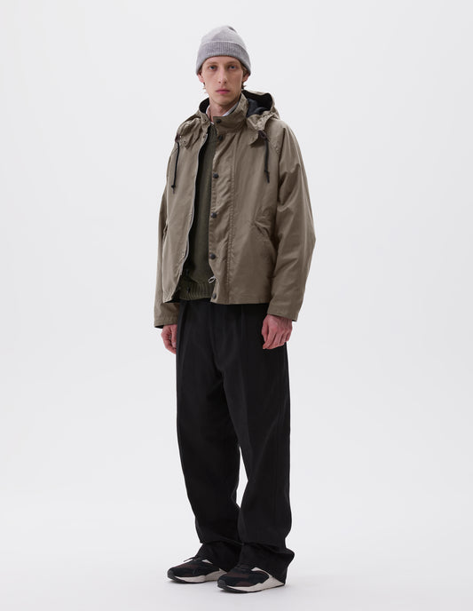 Full body image, front view of model wearing boxy dark beige jacket with stand collar and hood with wire adjustable peak, raglan sleeve, two-way zip fastening, press stud storm flap and wide storm welt side pockets. Styled over green jumper and white shirt with grey beanie, black trousers and black, brown and white trainers.