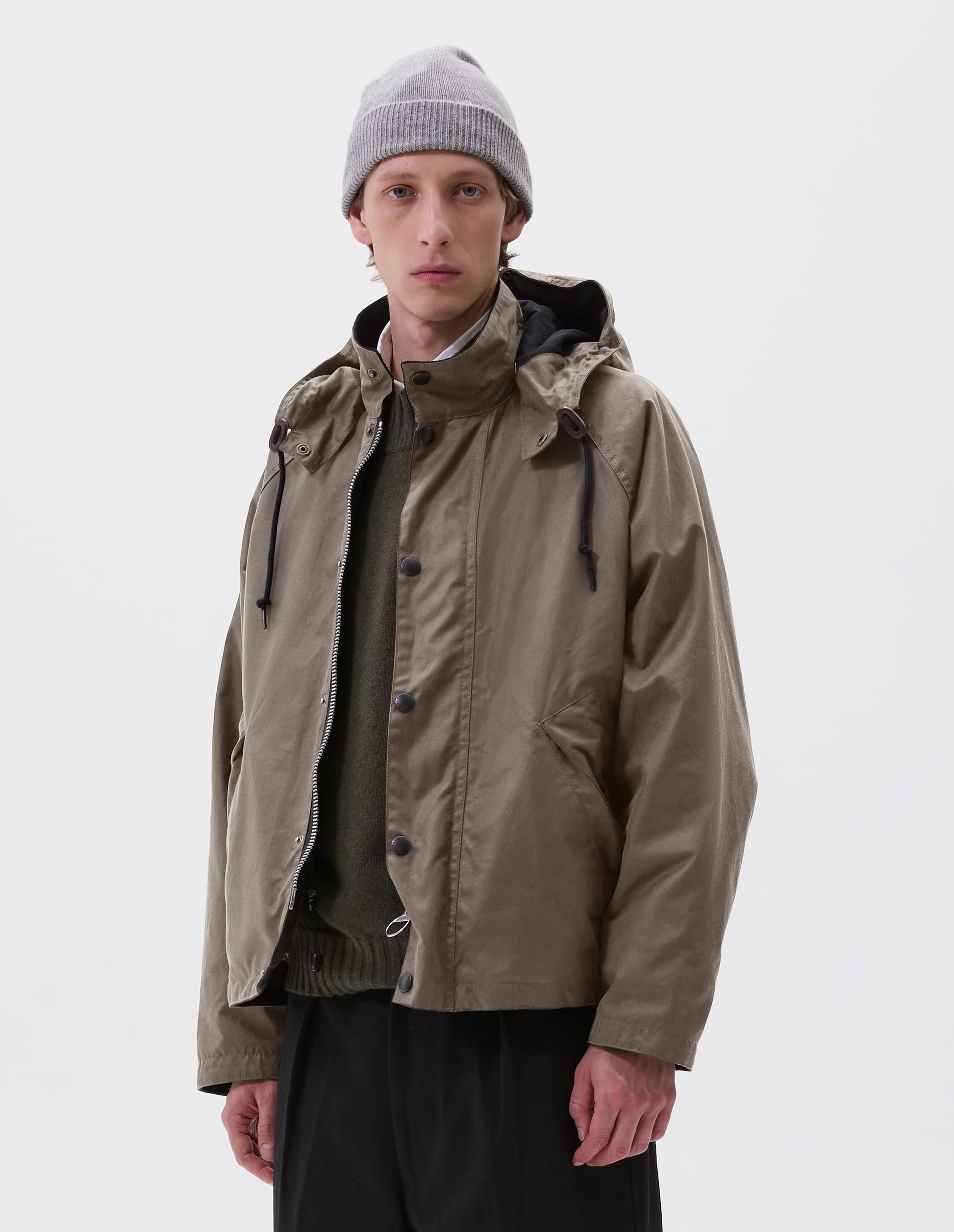 Cropped image, front view of model wearing boxy dark beige jacket with stand collar and hood with wire adjustable peak, raglan sleeve, two-way zip fastening, press stud storm flap and wide storm welt side pockets. Styled over green jumper with grey beanie and black trousers.