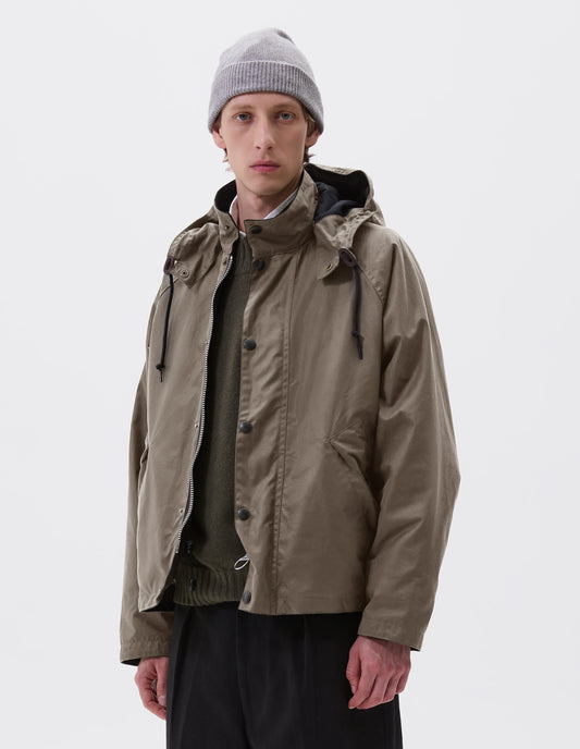 Cropped image, front view of model wearing boxy dark beige jacket with stand collar and hood with wire adjustable peak, raglan sleeve, two-way zip fastening, press stud storm flap and wide storm welt side pockets. Styled over green jumper with grey beanie and black trousers.