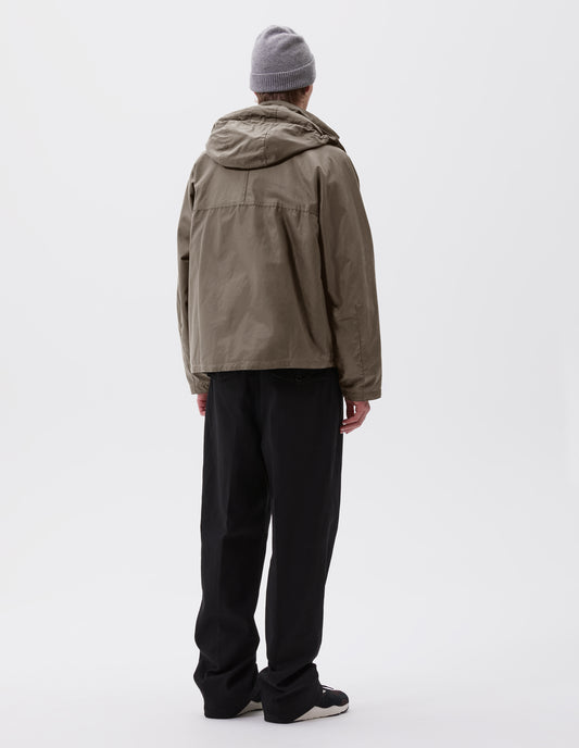 Full body image, back view of model wearing boxy dark beige jacket with raglan sleeve and hood with wire adjustable peak. Styled with grey beanie, black trousers and black, brown and white trainers.