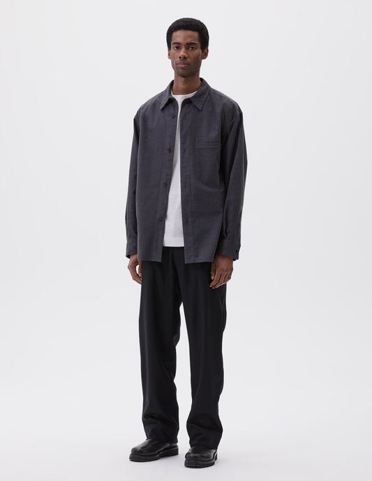 Full body image, front view of model wearing relaxed dark grey shirt with horn buttons and welt chest pocket. Styled over white t-shirt and black trousers and black leather boots.