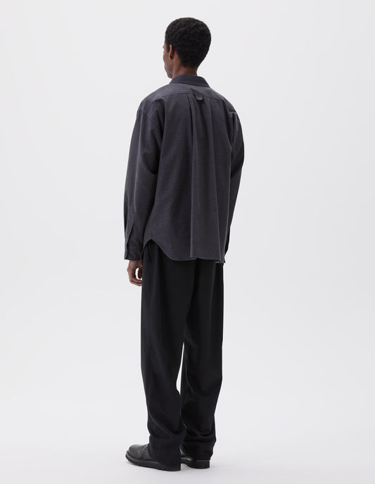 Full body image, back view of model wearing relaxed dark grey shirt with horn buttons on cuffs and wide box pleat and hanger loop detail on the back yoke. Styled with black trousers and black leather boots.
