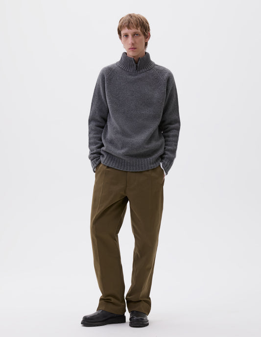 Full body image, front view of model wearing grey melange mock neck jumper with zip detailing, raglan sleeve and contrasting rib on neck, cuffs and hem. Styled with green trousers and black leather boots.