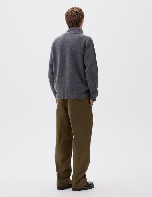 Full body image, back view of model wearing grey melange mock neck jumper with raglan sleeve and contrasting rib on neck, cuffs and hem. Styled with green trousers and black leather boots.