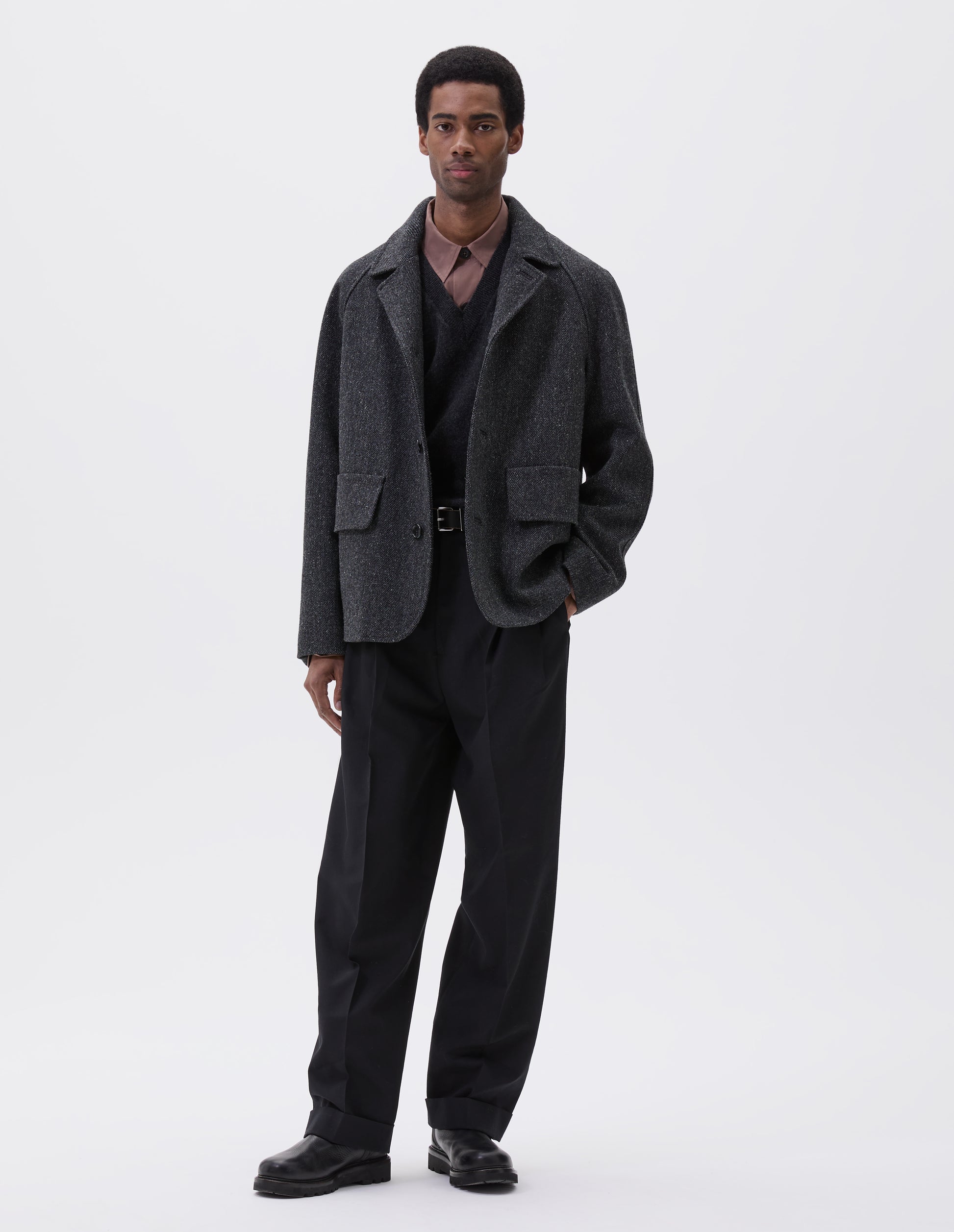 Full body image, front view of model wearing grey wool blazer with high collar fastening, deep flap welt pockets and subtle horn button detailing. Styled over pink shirt, grey v-neck sweater, black trousers and black leather boots. 