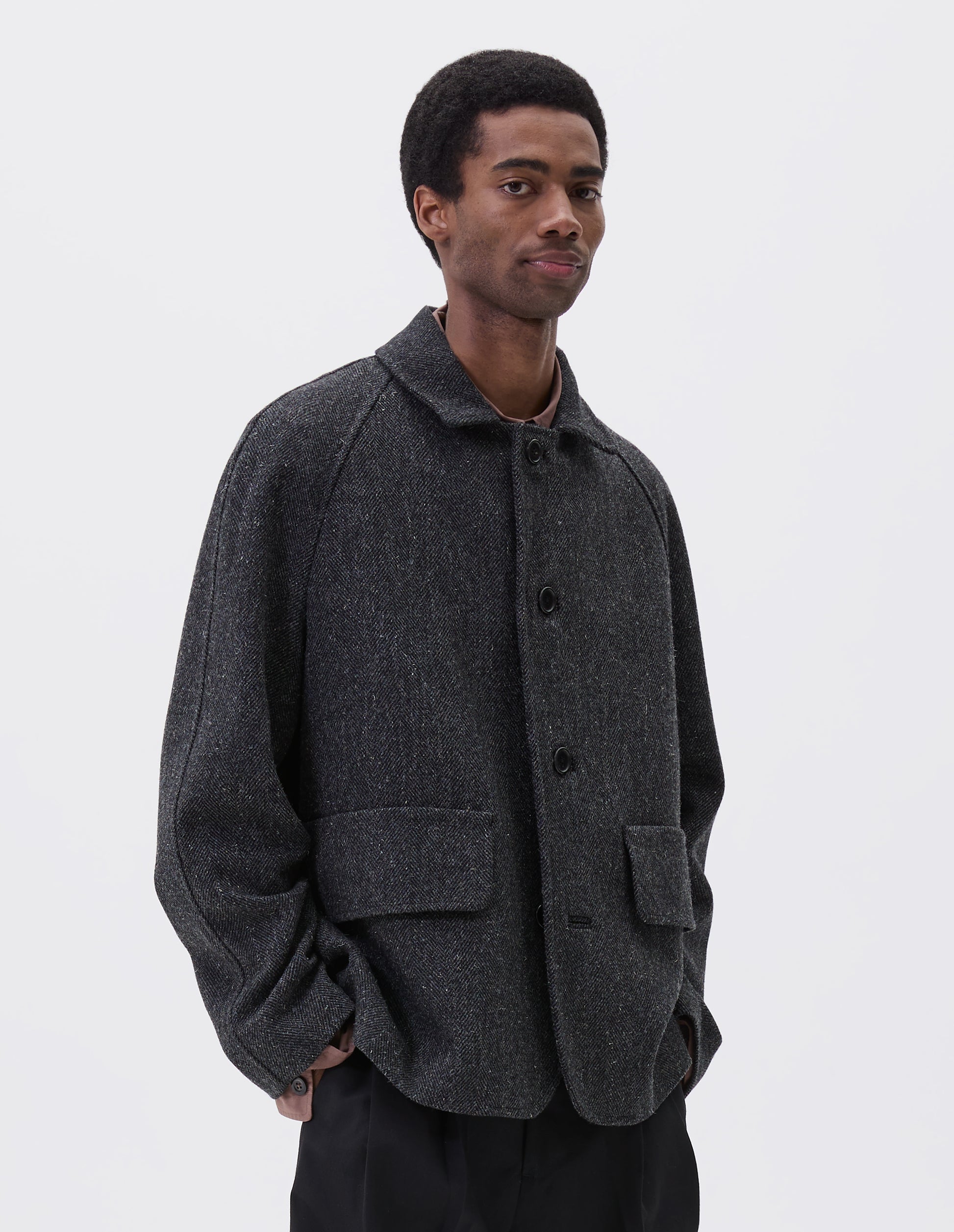 Cropped body image, front view of model wearing grey wool blazer with high collar fastening, deep flap welt pockets and subtle horn button detailing. Styled over pink shirt, with black trousers.