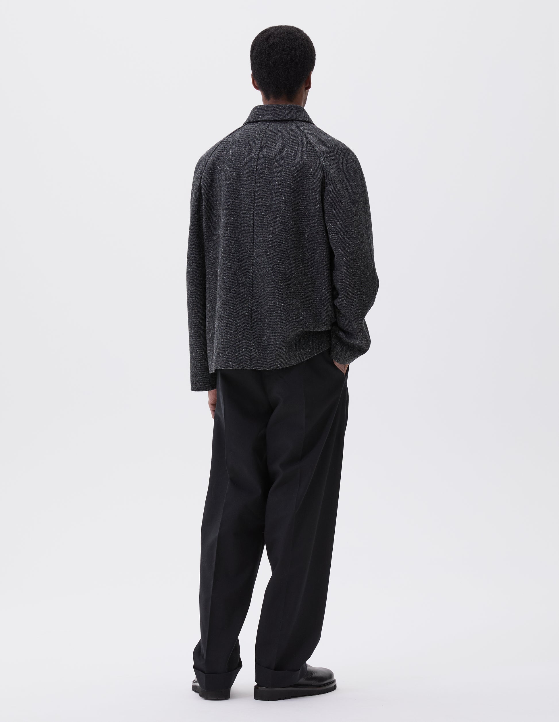 Full body image, back view of model wearing grey wool blazer with high collar fastening and raglan sleeve Styled with black trousers and black leather boots. 