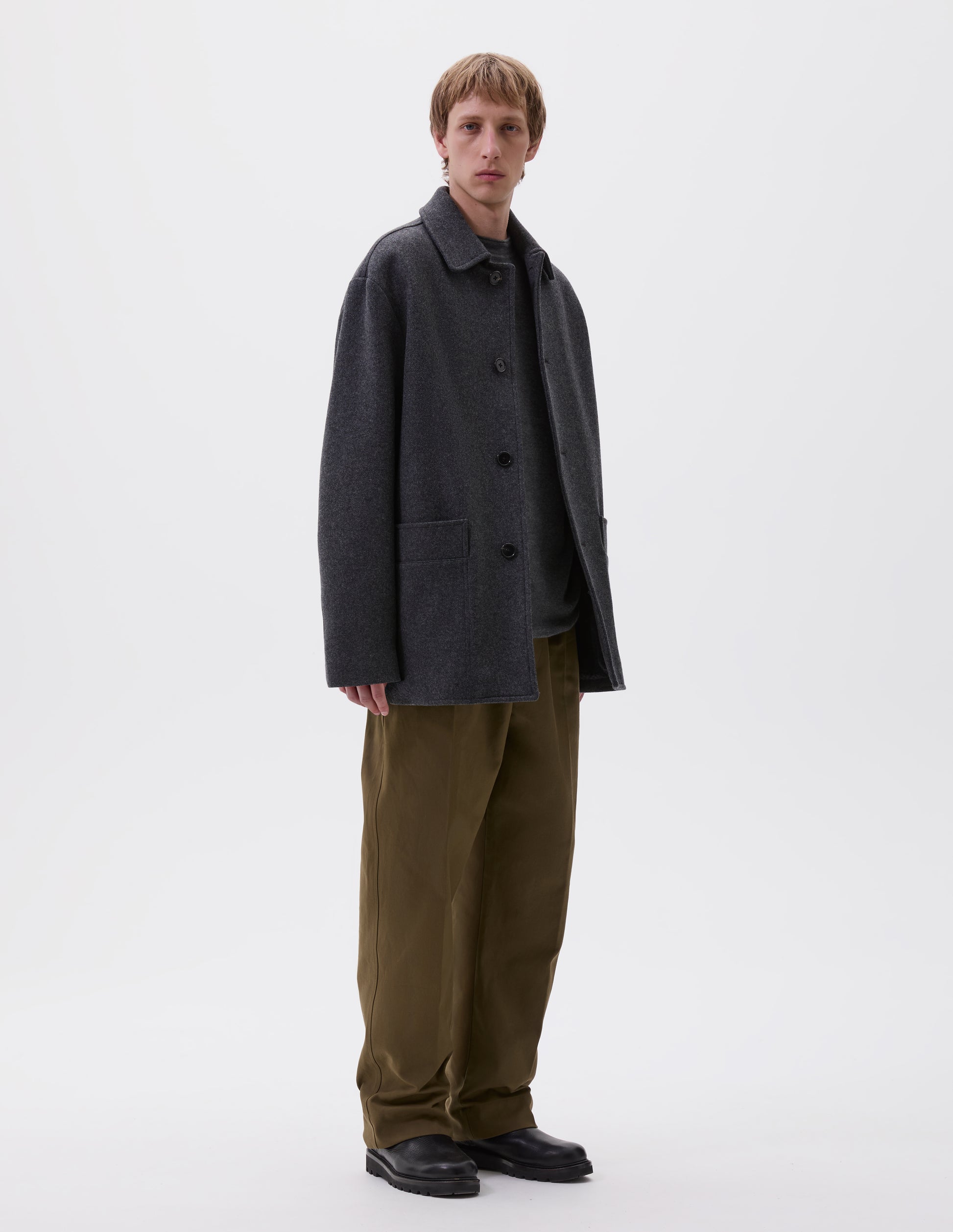 Full body image, front view of model wearing charcoal grey wool coat with deep relaxed welt pockets and stitch through detailing. Finished with horn button closure. Styled with grey jumper, green trousers and black leather boots.