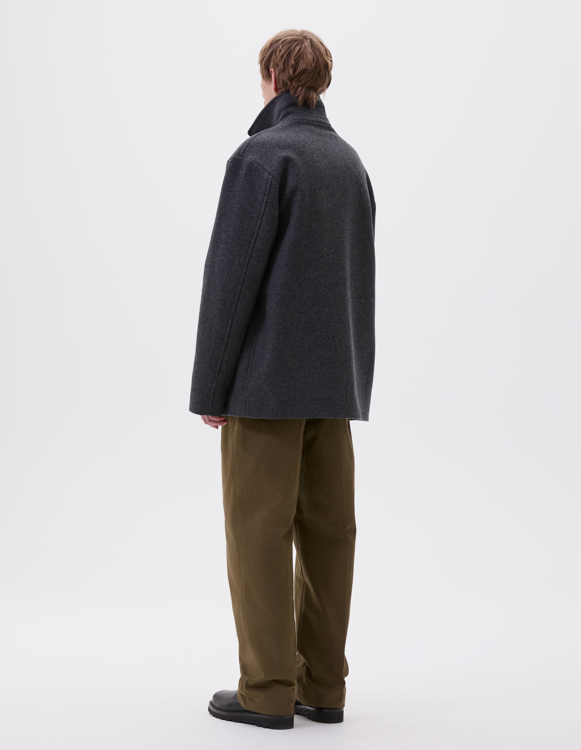 Full body image, back view of model wearing charcoal grey wool coat with contrast wool cotton fleece lining, with deep relaxed welt pockets and stitch through detailing. Styled with green trousers and black leather boots. 